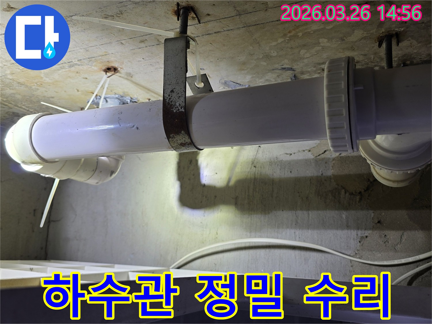 콘크리트 천장에 고정된-하얀색 하수관의 연결 부위와-녹슨 지지대를 엔지니어가-정밀하게 점검하는 모습으로-수리 전 진단 과정을 보여줍니다