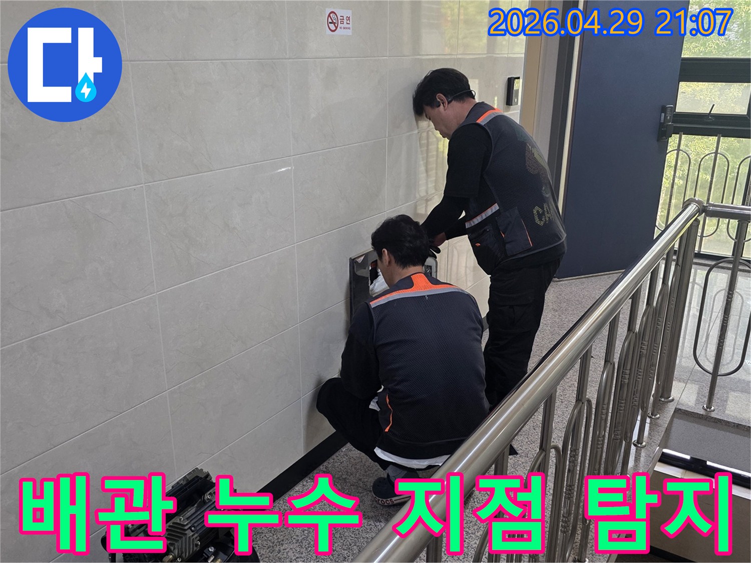 두 명의 전문 기술자가 건물 벽면 내부의 배관 누수 의심 지점을 첨단 장비로 정밀하게 탐지하고 있습니다-정확한 원인 파악을 위한 집중적인 작업입니다.