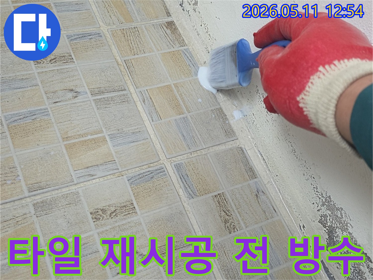 붉은 장갑을 낀 작업자가-베란다 타일 재시공 전-벽면 하단과 바닥 경계에-방수액을 붓으로 꼼꼼히 바르며-밑작업을 진행하고 있습니다