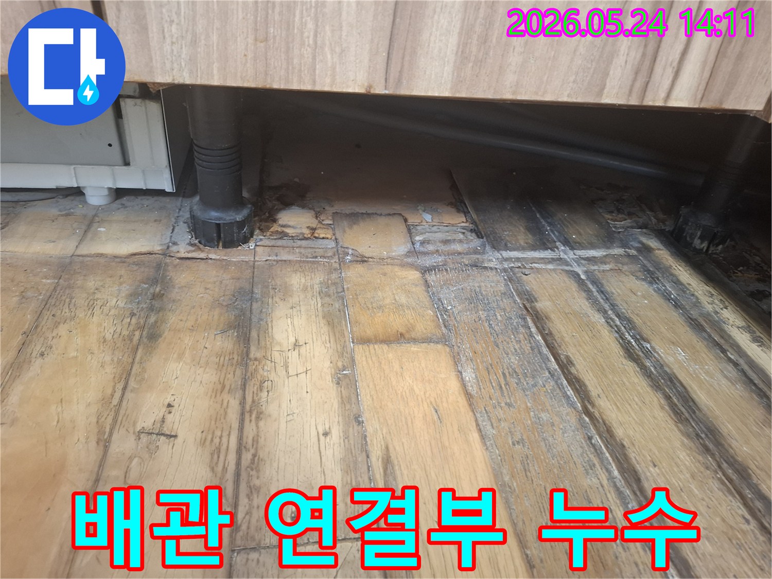 물에 젖어 어둡게 변색되고 부식된 나무 바닥과-그 옆에 위치한 검은색 배관 연결부가 보입니다
