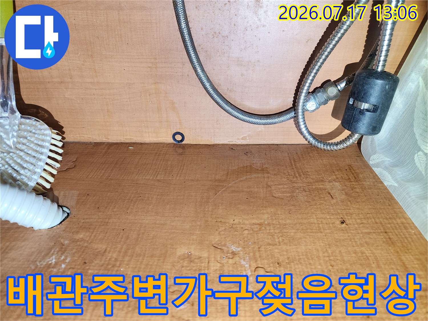 물에 젖어 변색된 주방 하부장의 목재 벽면-금속제 배관과 흰색 자바라 호스가 설치된 장면