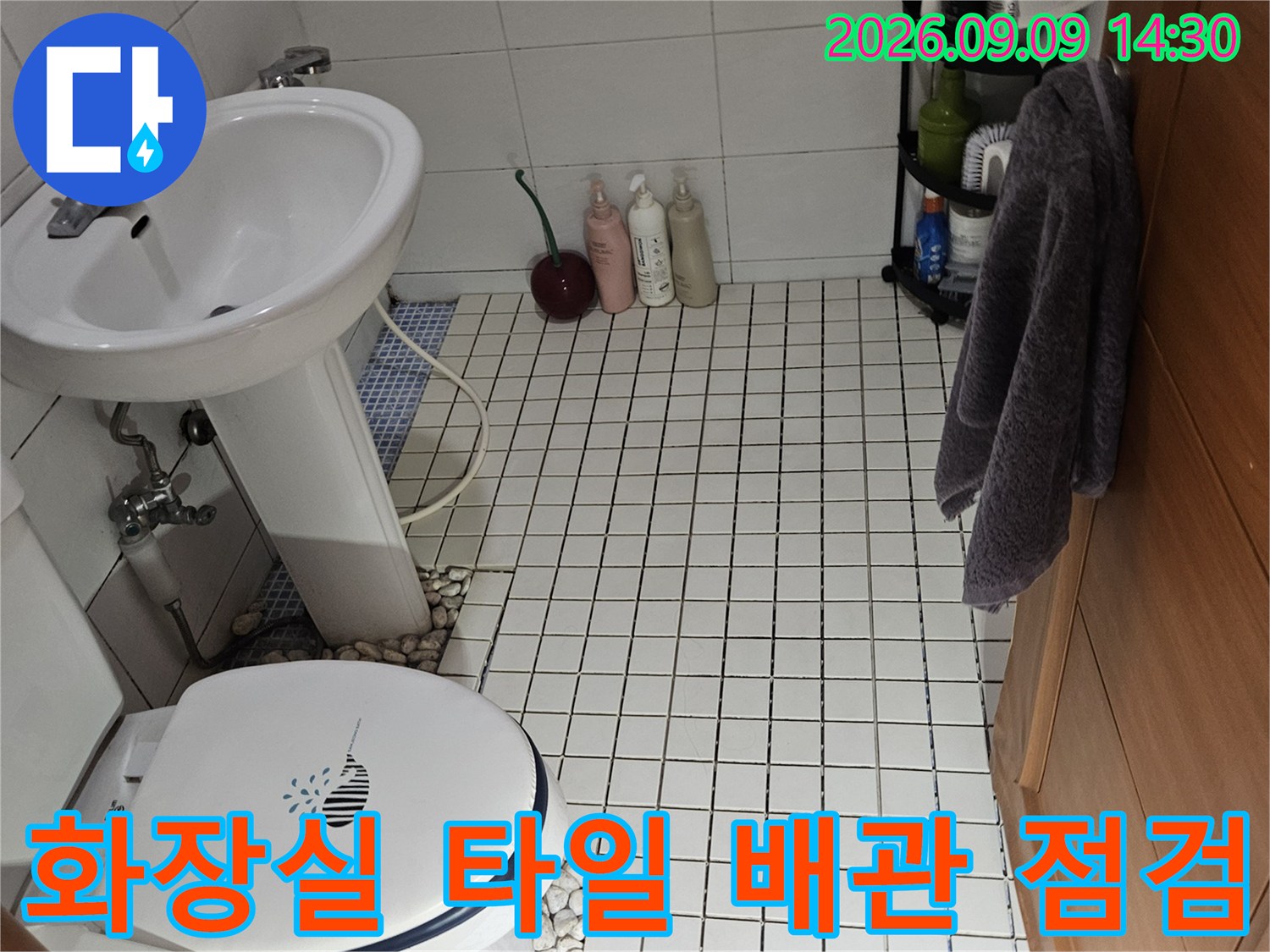 흰색 타일로 마감된 욕실 벽면과 세면대 하부 배관의 연결 상태를 촬영하였습니다.