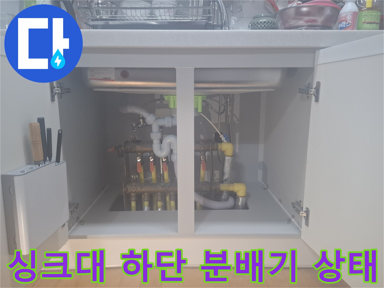 주방 싱크대 하부장에 위치한 난방 분배기와 배관 연결 부위 모습입니다.