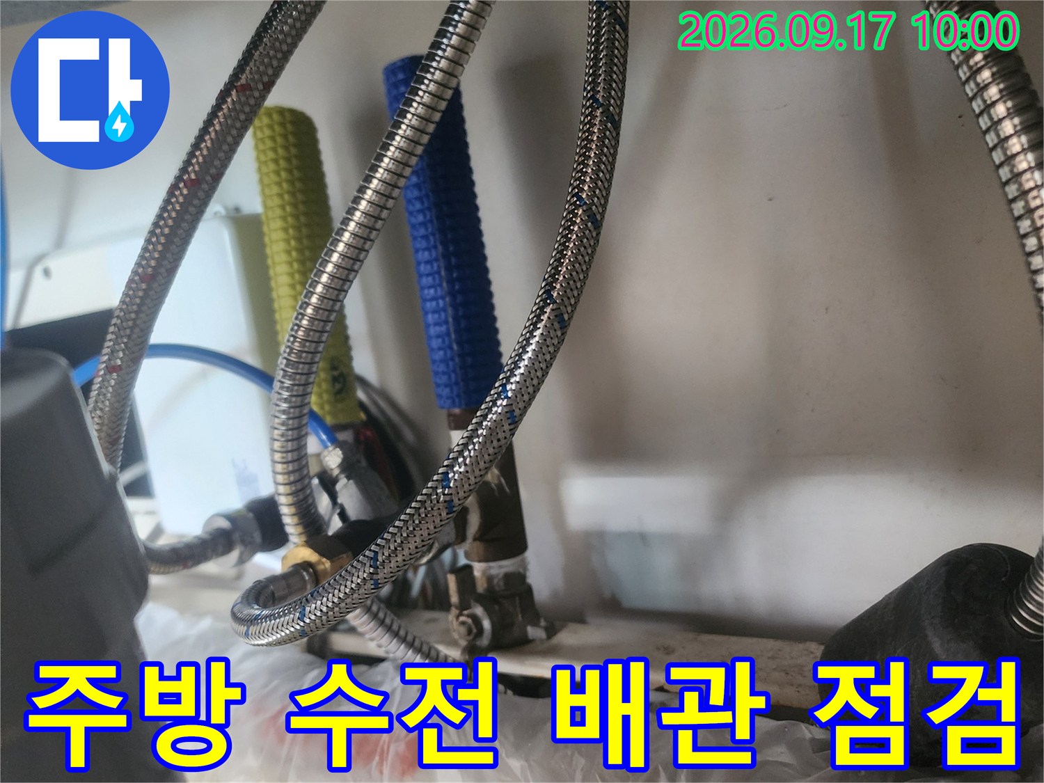 은색 꼬임 호스와 파란색 배관이 보이는 싱크대 하부 내부입니다.
